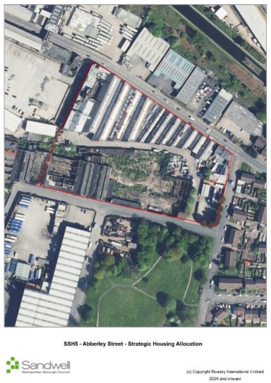 Aerial view of an urban industrial area with a red boundary outlining a section that includes large warehouses, elongated buildings, and surrounding roads. Adjacent green open space is visible at the bottom.