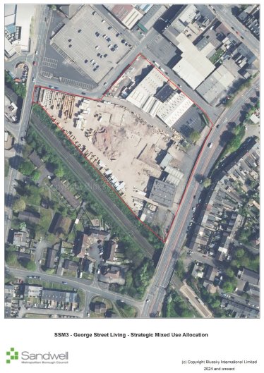 Aerial view of a construction site within a triangular boundary outlined in red, surrounded by buildings, roads, and green spaces. The site has some existing structures and cleared areas, with nearby parking lots and residential houses.