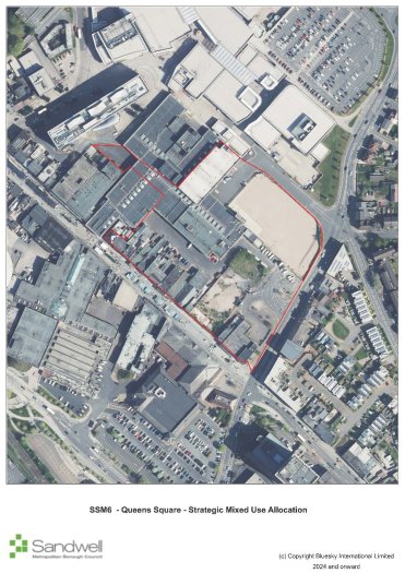 Aerial view of a city block with buildings, parking lots, and roads, enclosed by a red line indicating a designated area.